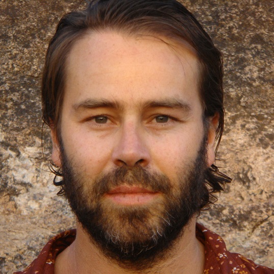 Photo of Shaun Parker, Shaun has a short beard and moustache, freckles on his nose, and has lightly tanned skin, he wears a red wine colour shirt on which is barely visible. He stands infront of a textured stone wall. He has a gentle smile and friendly eyes, his hair is brown and long-ish above the shoulders